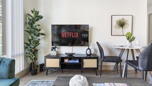 Moderni olohuone, jossa on seinälle asennettu taulutelevisio, jossa näkyy teksti ”NETFLIX HIT”, musta viihdekeskus, pieni ruokailutila, jossa on pieni pöytä ja tuolit, sekä suuri ruukussa oleva kasvi.