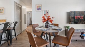 Un espace repas moderne avec une table ronde, des chaises marron et un vase rempli de fleurs rouges. Les murs sont ornés d’œuvres d’art encadrées et un téléviseur affiche le logo de Netflix.