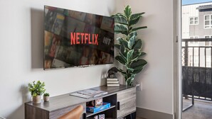 Un salon avec un téléviseur fixé au mur qui affiche le logo de Netflix, un meuble en bois contenant des jeux de société et une grande plante verte.