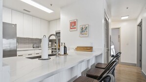 Une cuisine moderne avec des meubles blancs, un plan de travail en marbre et un four encastré. On y trouve deux tabourets de bar et une planche à découper en bois posée sur le plan de travail.