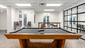 A pool table in a room with white walls, a wooden floor, and large windows.