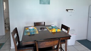 A dining area with a table set for two, chairs, a trash bin, and a wall-mounted light switch. - Charming 1-bedroom apartment in beautiful Black Point with amazing views (Black Point)