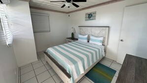 A bedroom with a bed, a ceiling fan, a nightstand, a lamp, a window with blinds, and a door. - Charming 1-bedroom apartment in beautiful Black Point with amazing views (Black Point)