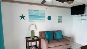 A room with a ceiling fan, a painting of the sea, a star decoration, a clock, a 'No Smoking' sign, a television, a sofa with teal cushions, a small table with a lamp, and a magazine on the table. - Charming 1-bedroom apartment in beautiful Black Point with amazing views (Black Point)