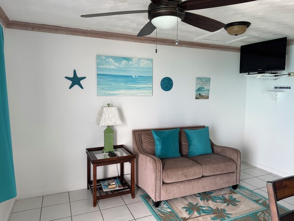 A room with a ceiling fan, a painting of the sea, a star decoration, a clock, a 'No Smoking' sign, a television, a sofa with teal cushions, a small table with a lamp, and a magazine on the table. - Charming 1-bedroom apartment in beautiful Black Point with amazing views (Black Point)
