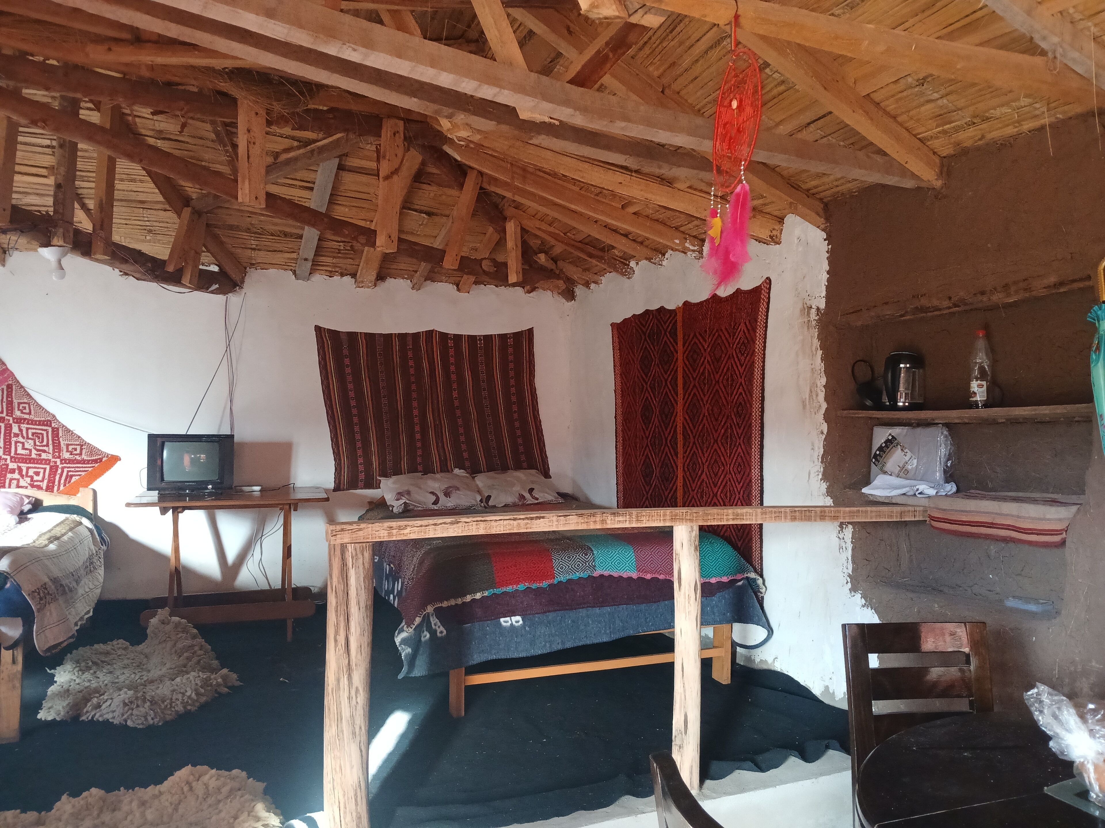 Una habitación rústica con techo de madera, una cama con colcha colorida, un televisor y una repisa con un hervidor y una botella de plástico.