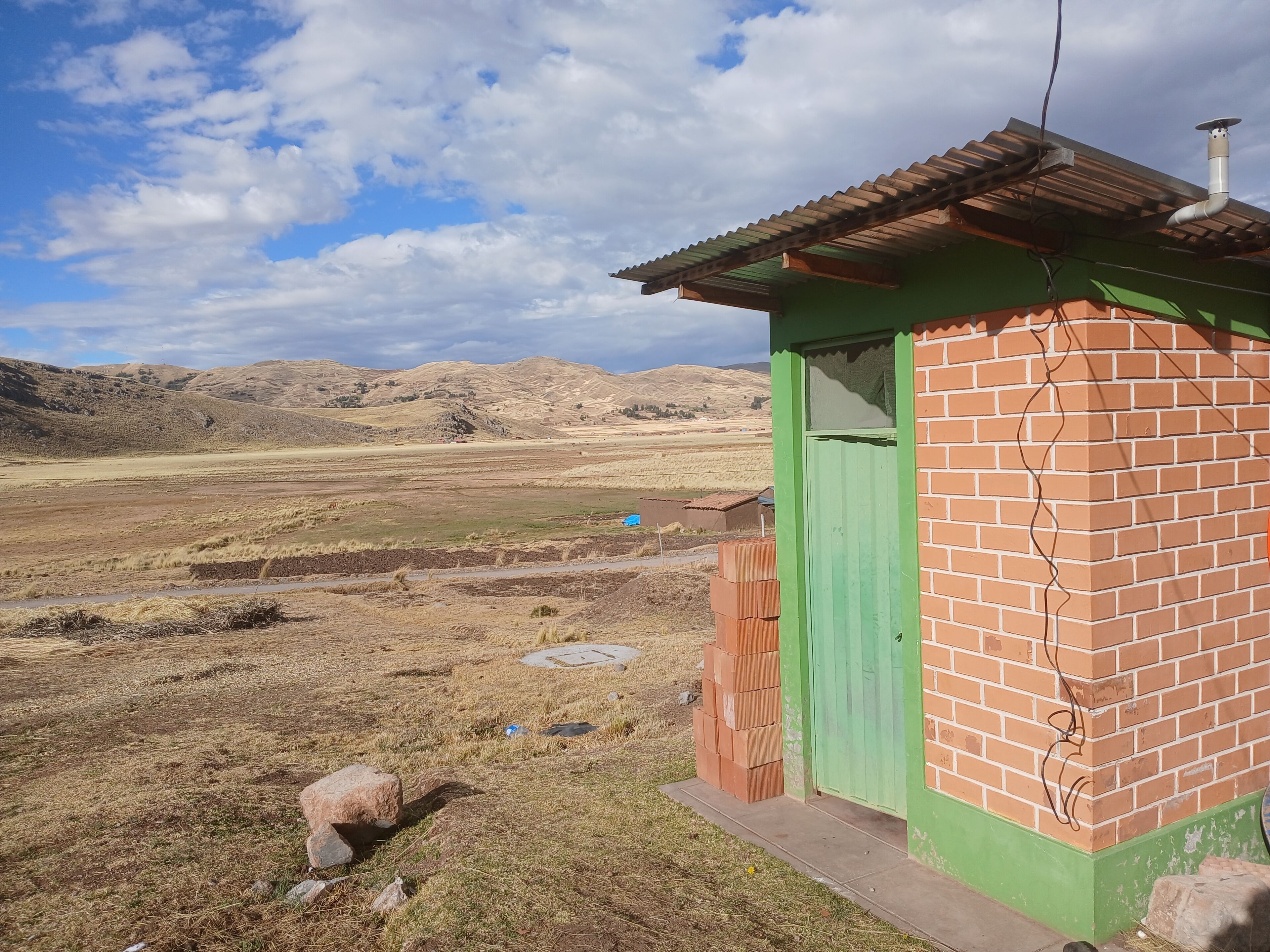 Um pequeno edifício de tijolos com uma porta verde, localizado em uma paisagem rural com colinas ao fundo.