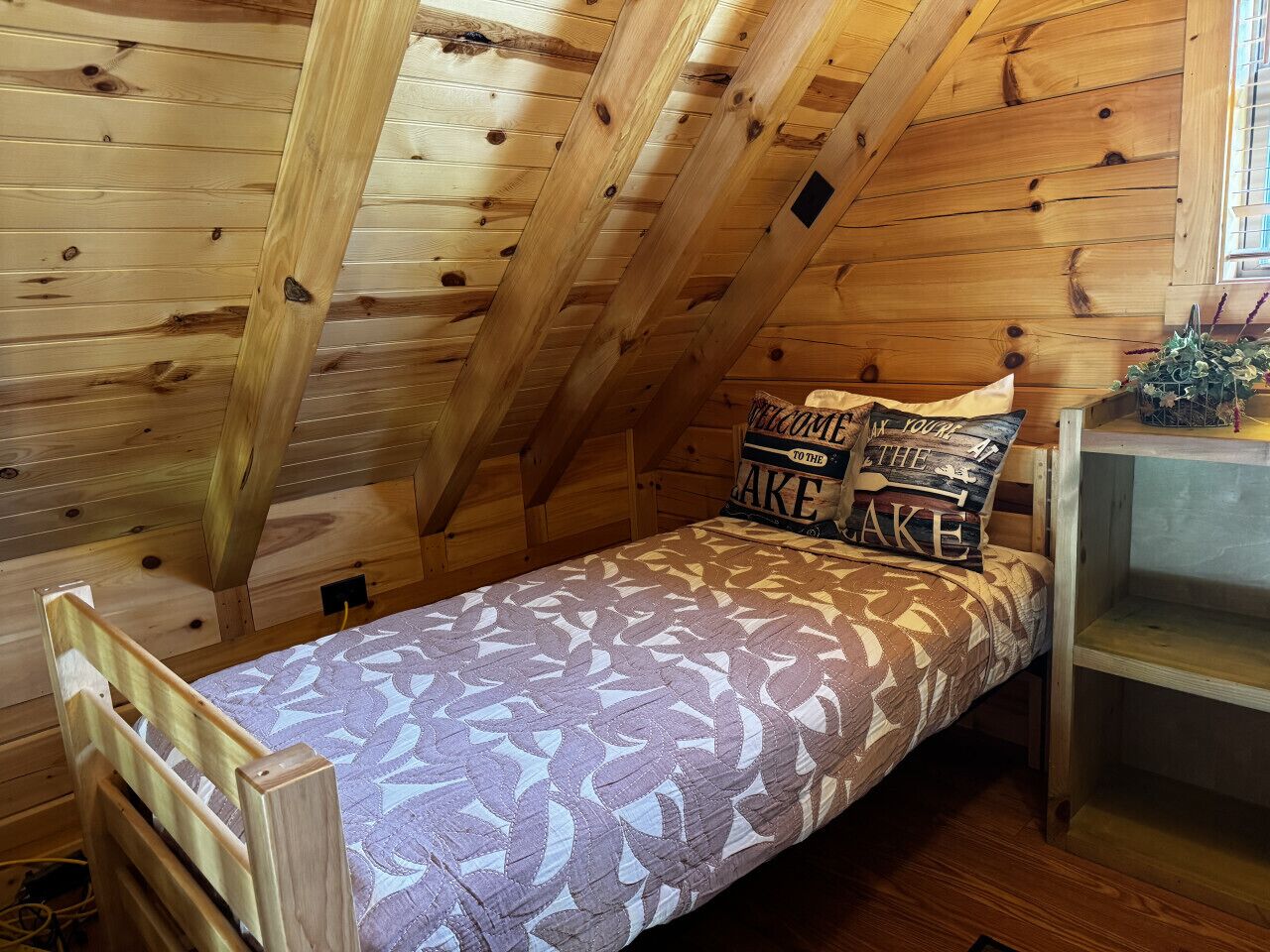 A small, cozy room with a sloped ceiling, wooden walls, a single bed with patterned bedding, and a small shelf with decorative items.