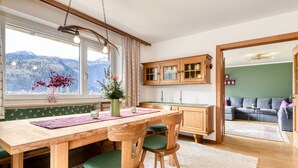 Un coin repas avec une table et des chaises en bois, une vue sur les montagnes à travers de grandes fenêtres et un canapé vert dans le salon attenant.
