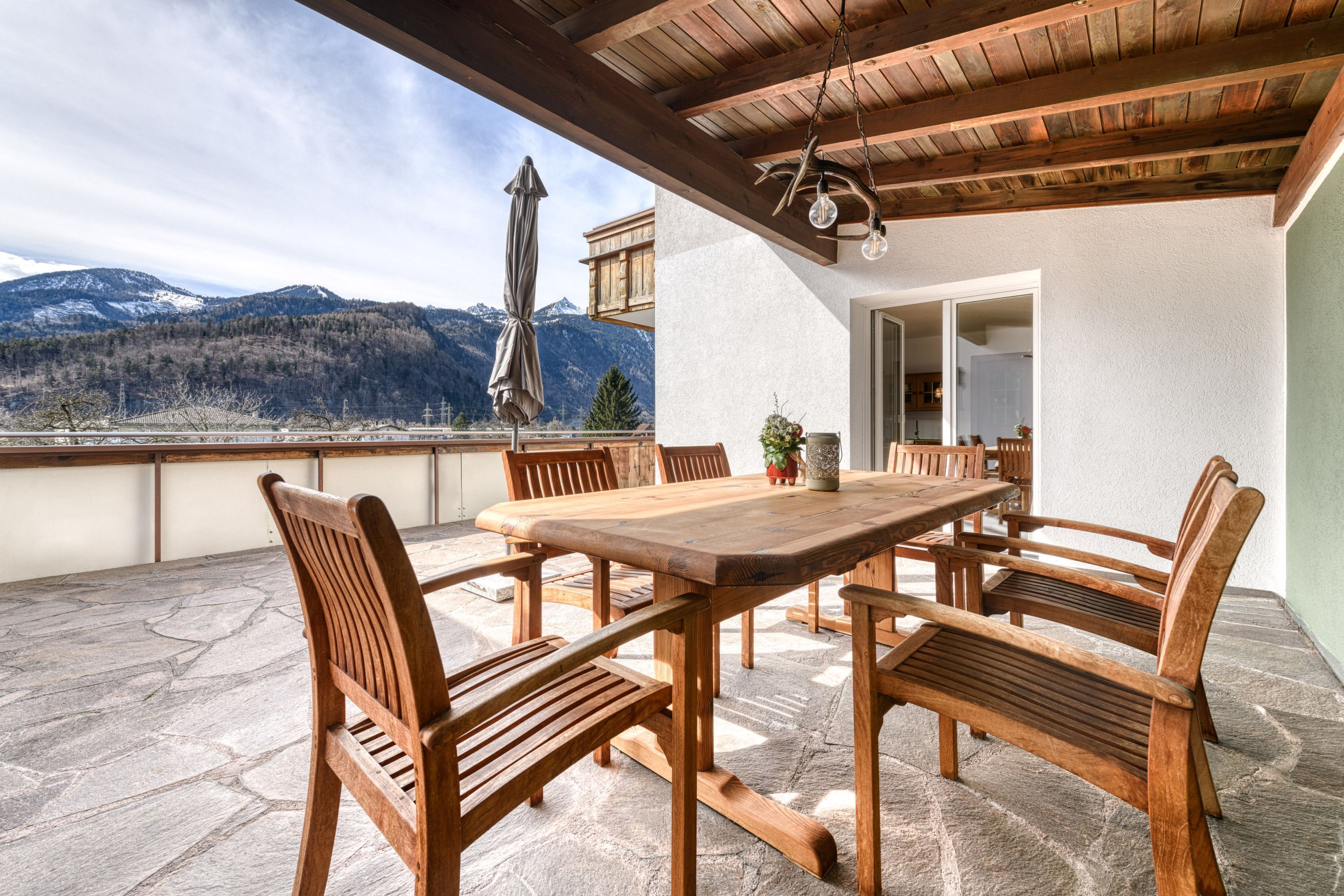 En udendørs terrasse med et træbord og stole, stenbelagt gulv og udsigt til bjerge.