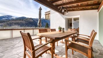 An outdoor patio with a wooden table and chairs, stone flooring, and a view of mountains.