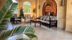 A spacious living area with a sofa, chairs, and a coffee table. There are two windows with arched tops, a decorative wall, and a large plant in the foreground. - Fully furnished 02 BR with sea view roof top (Hurghada)