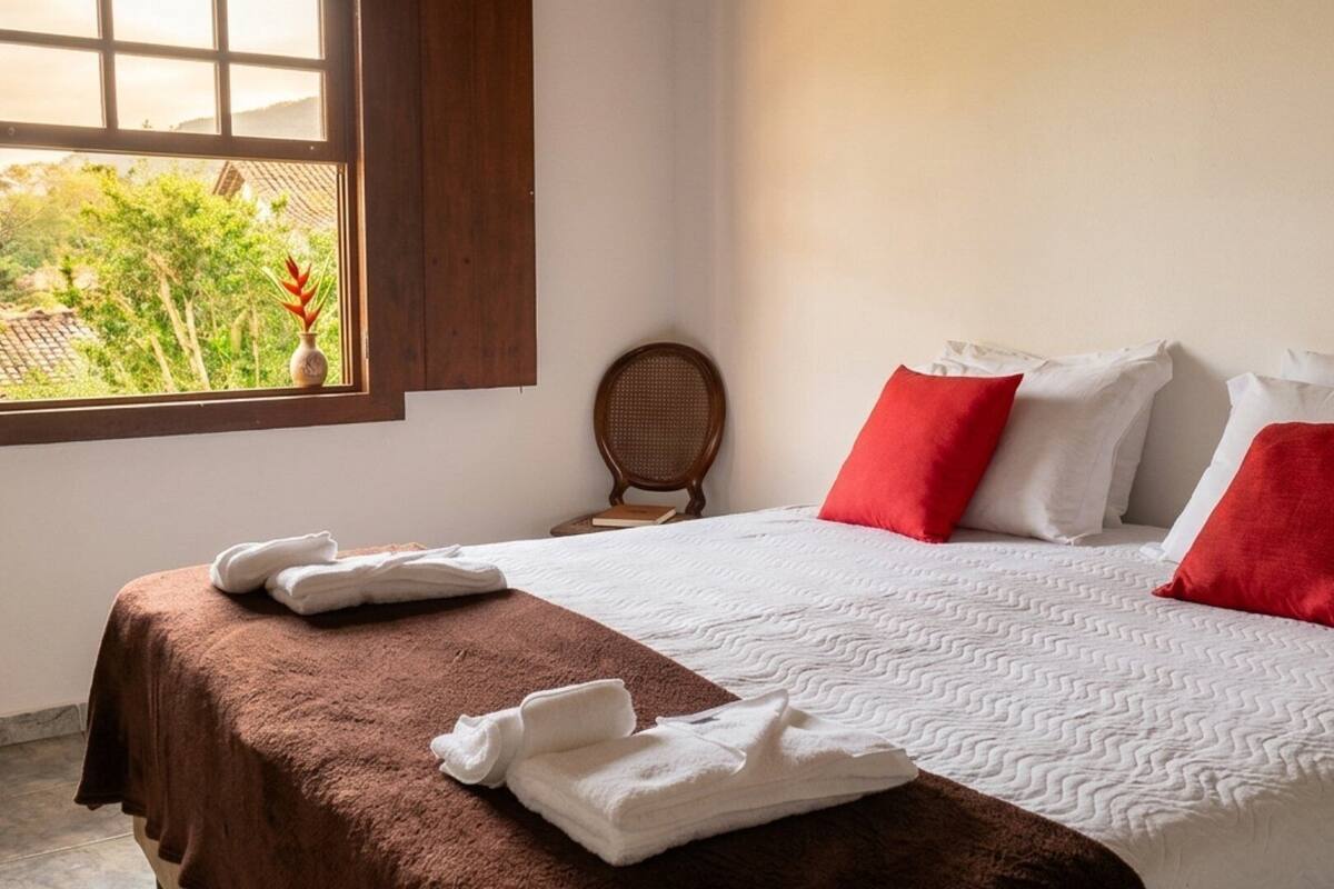 A neatly made bed with white and red pillows, a brown headboard, and a window with a view of greenery.