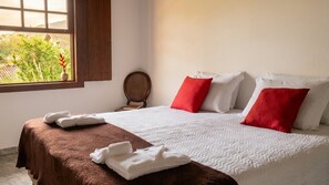 A neatly made bed with white and red pillows, a brown headboard, and a window with a view of greenery. - Refúgio Tiradentes (Tiradentes)