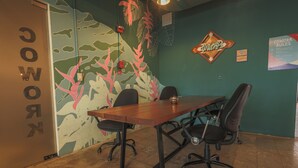 Ein Konferenzraum mit einem Holztisch und vier Stühlen, einer grünen Wand mit einem Blatt-Wandbild und einem Plakat mit der Aufschrift 'Cowork Rules'.
