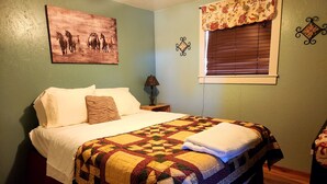 Una cama bien hecha con colcha, una mesita de noche con lámpara, un valance floral en la ventana y un cuadro con tema de caballos en la pared.