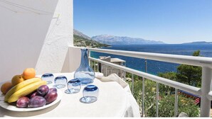 A balcony with a table set with fruits and glasses, overlooking a coastal view with mountains. - Beautiful apartment in Omis with lake view (Omis)