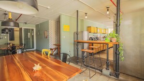 Un comedor amplio con una mesa de madera, sillas y una lámpara colgante grande. Se observa una zona de cocina con armarios y un refrigerador al fondo.