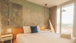 Habitación de hotel con una cama grande, cabecera de madera, mesita de noche, lámpara y vista a la ciudad por la ventana.