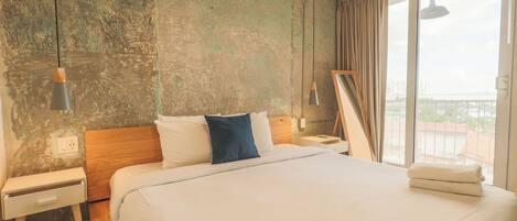 A hotel room with a large bed, a wooden headboard, a nightstand, a bedside table, a lamp, and a view of the city through the window.