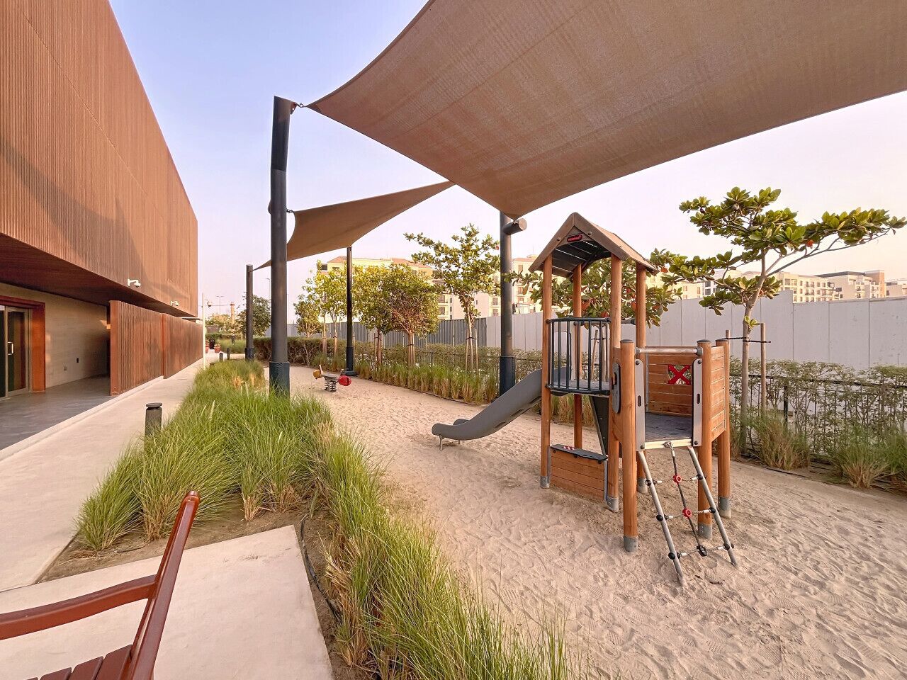 Un'area giochi con scivolo e strutture per arrampicarsi, circondata da sabbia e vegetazione, con un edificio moderno nelle vicinanze.