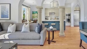 A spacious living area with a grey sofa, a small round table, and a blue armchair. There is a kitchen in the background with white cabinets and a blue countertop.