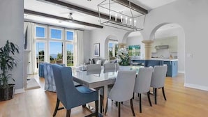 A spacious dining area with a modern chandelier, a large window with a view, and a kitchen in the background.