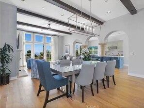 Enjoy meals in this stylish dining area with a large table and six chairs, surrounded by a high ceiling and large windows offering views of the surrounding landscape.