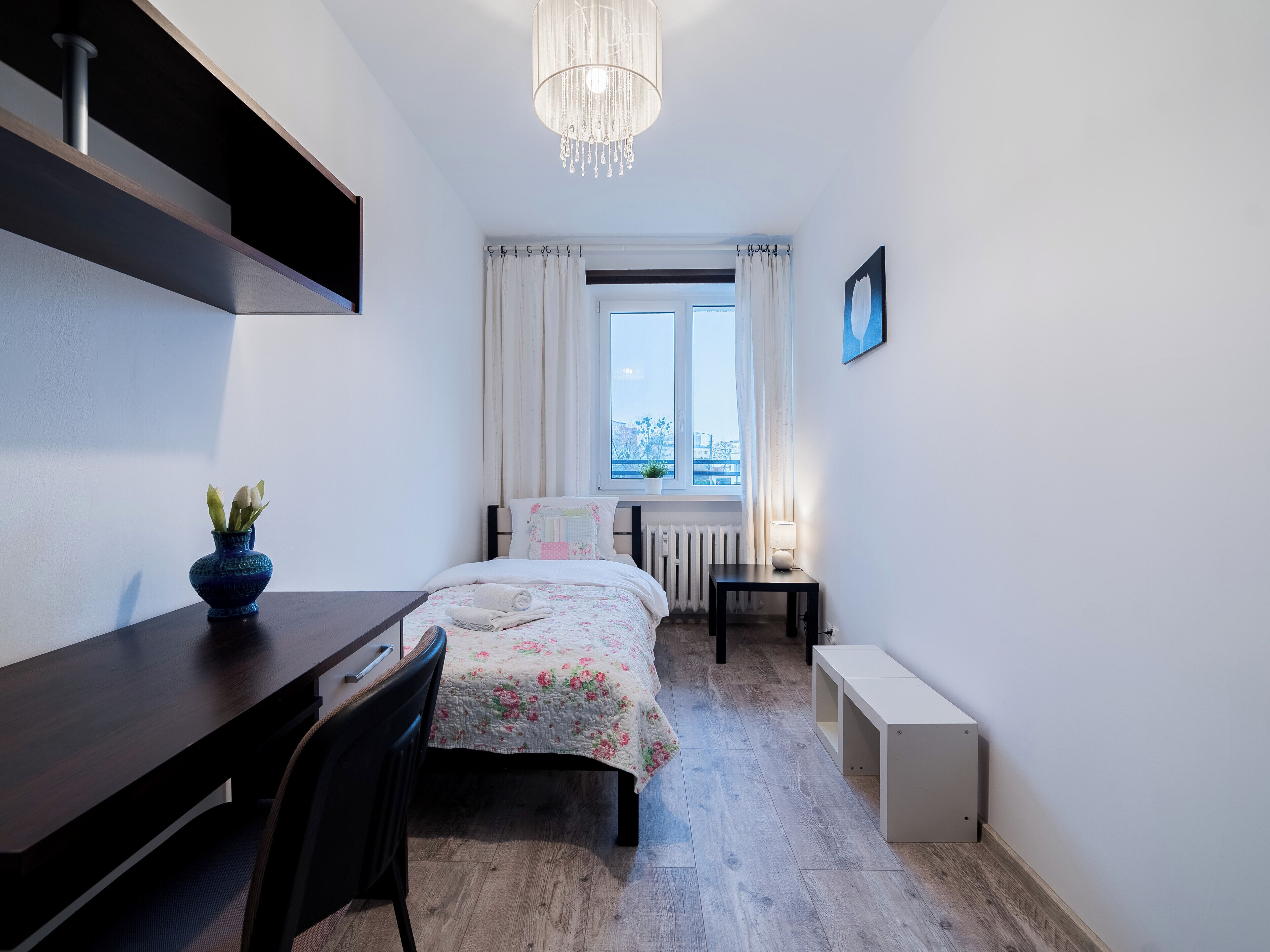 Une chambre à coucher de taille réduite, comprenant un lit, un bureau, une chaise, une petite table, un radiateur, une fenêtre avec des rideaux et un luminaire suspendu.