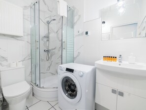 A modern bathroom with a glass-enclosed shower, a built-in washing machine, and a white sink with toiletries.