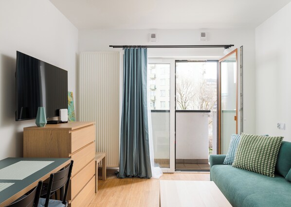 A modern living room with a flat-screen TV, wooden furniture, a green sofa, and a sliding glass door leading to a balcony.