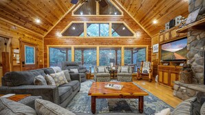 A spacious living room with a high ceiling, wooden walls, and large windows offering a view of the outdoors. There is a fireplace with a stone surround and a television mounted on the wall.