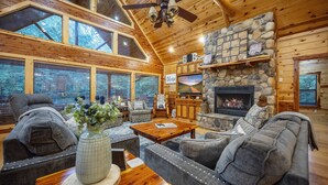 A cozy log cabin interior with a stone fireplace, wooden furniture, and large windows.
