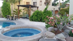 A swimming pool with a hot tub, surrounded by rocks and plants, with outdoor seating and a pergola.