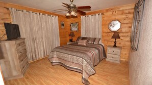 A bedroom with a wooden floor, a bed with patterned bedding, a ceiling fan, two bedside tables with lamps, a dresser, and curtains.