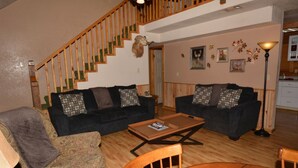 A living room with two dark-colored sofas, a wooden coffee table, a patterned armchair, a wooden dining table, and a staircase with a wooden railing.