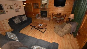A cozy log cabin interior with a sofa, armchair, fireplace, television, and a dining table.