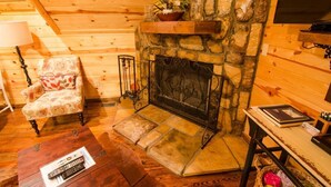 A cozy log cabin interior with a stone fireplace, a patterned armchair, and a wooden table with a remote control. - Squirrel's Nest is a perfect two-bedroom hideaway with tree top views! (Broken Bow)