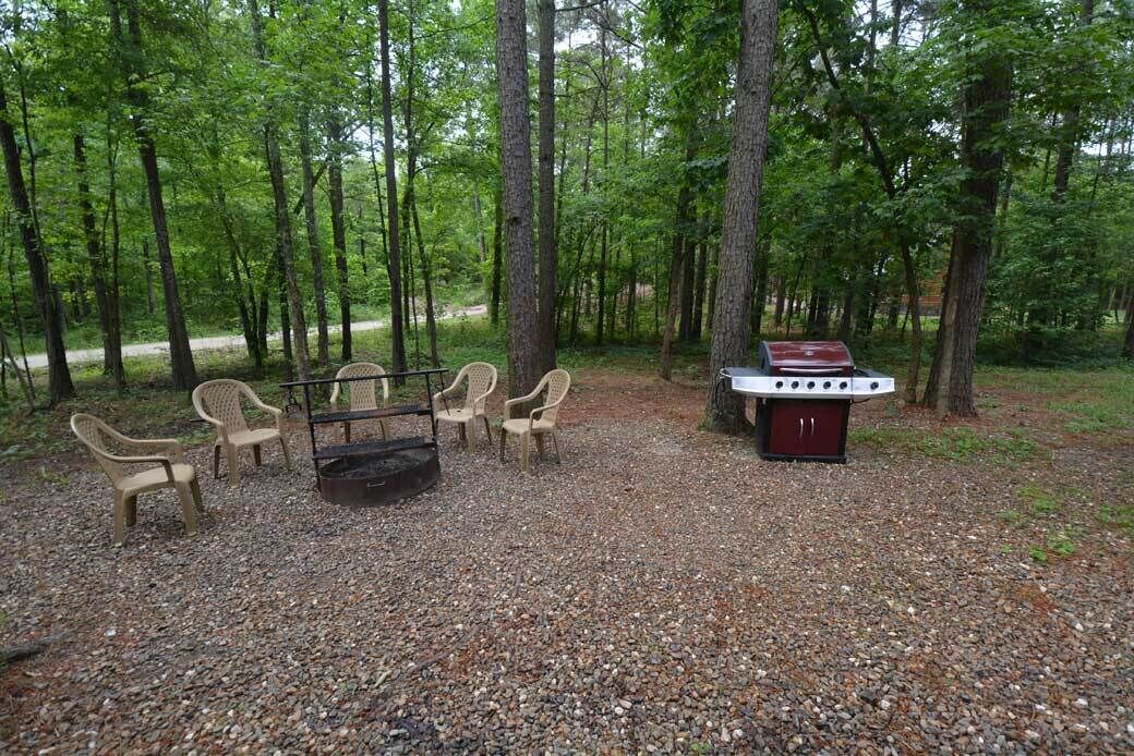 A campsite with a grill, chairs, and a fire pit surrounded by trees.
