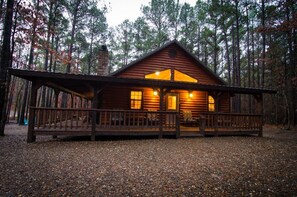 Una cabaña de troncos con porche y chimenea, rodeada de un bosque.