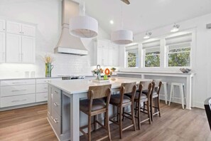 A modern kitchen with white cabinetry, a large island with bar stools, pendant lighting, and a view through large windows. - Persimmons 9 by Brunswick Vacation Rentals: 8BR w/ Elevator, Pool & Beach Access (Oak Island)