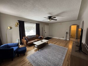 A living room with a brown leather sofa, a blue armchair, a coffee table, and a patterned rug. There is a ceiling fan, a floor lamp, and a door leading to another room. - Newly remodeled 2-bedroom house with WiFi, conveniently located in Great Falls (Great Falls)