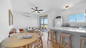 Una cocina moderna con una isla grande, una zona de comedor con sillas de madera y vistas a la playa a través de amplias ventanas.