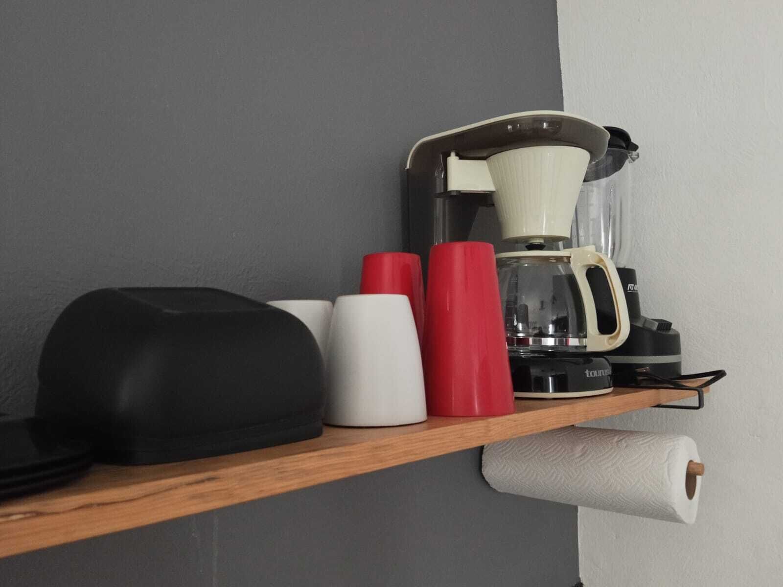 Eine Kaffeemaschine auf einem Regal, zusammen mit einer Toilettenpapierrolle, einem schwarzen Gegenstand und zwei roten Tassen.