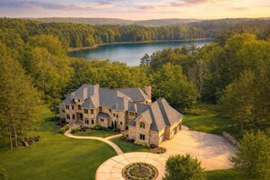 A large stone house with multiple chimneys, surrounded by a well-maintained lawn and a lake. - Gray Lake Villa | Sleeps 22 · Theater · Sauna · Lake (Gillett)