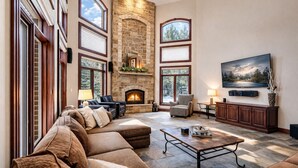 Un salón espacioso con chimenea de piedra, ventanas grandes, un sofá modular, una mesa de centro y un televisor de pantalla plana montado en la pared.