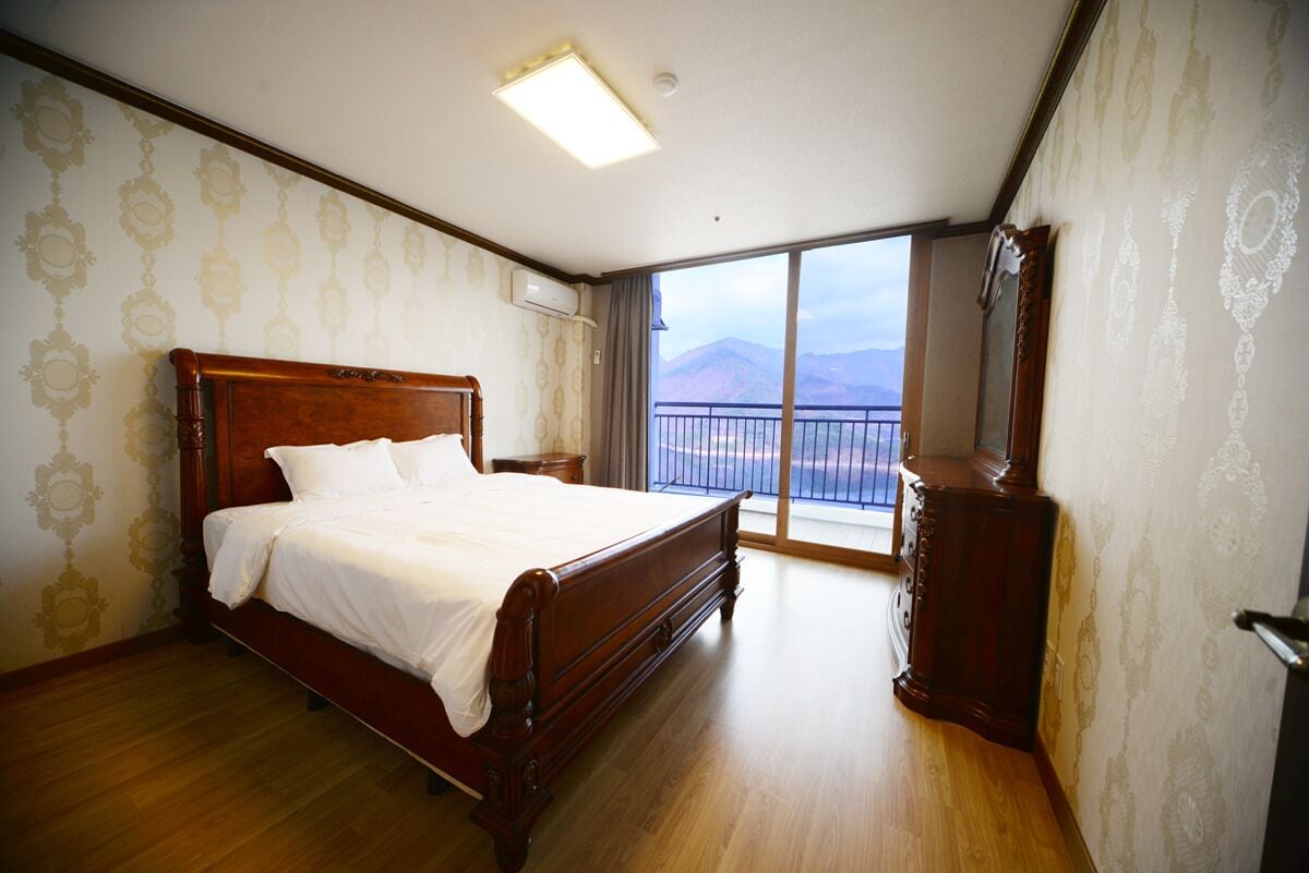 A bedroom with a large bed, a wooden dresser, and a balcony with a mountain view.