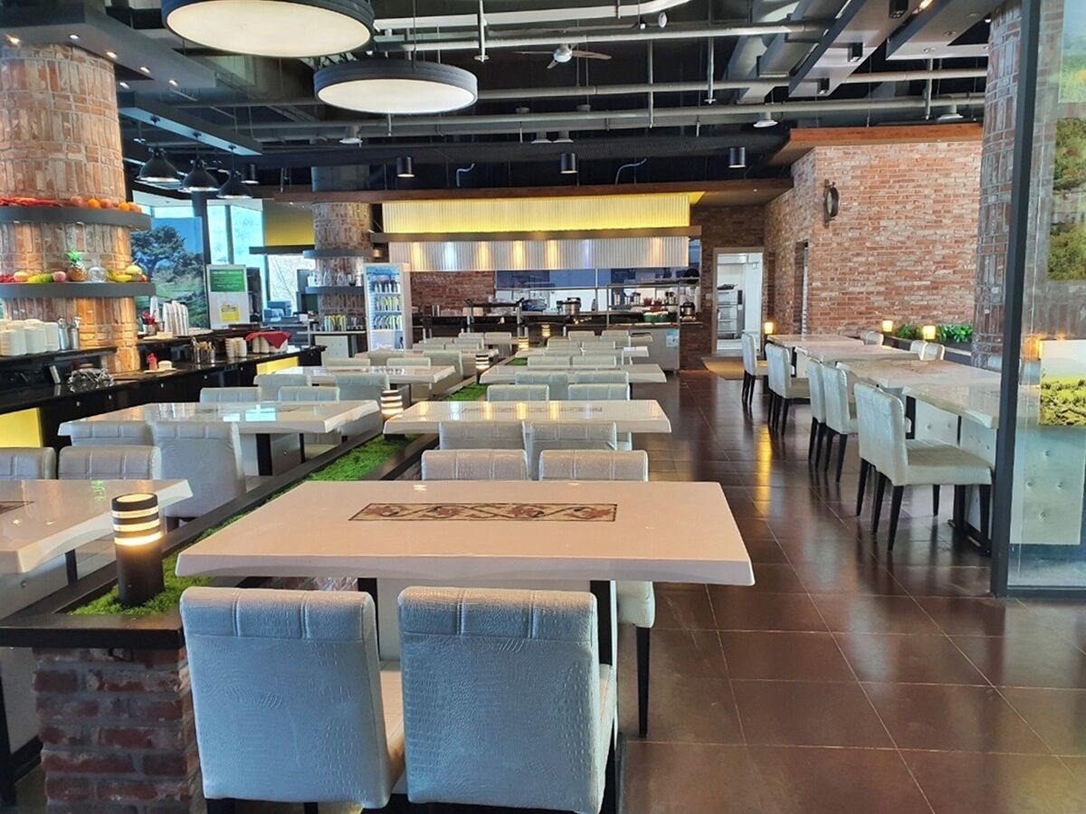 Une cafétéria spacieuse avec de nombreuses tables et chaises, un mur en briques et une cuisine moderne en arrière-plan.