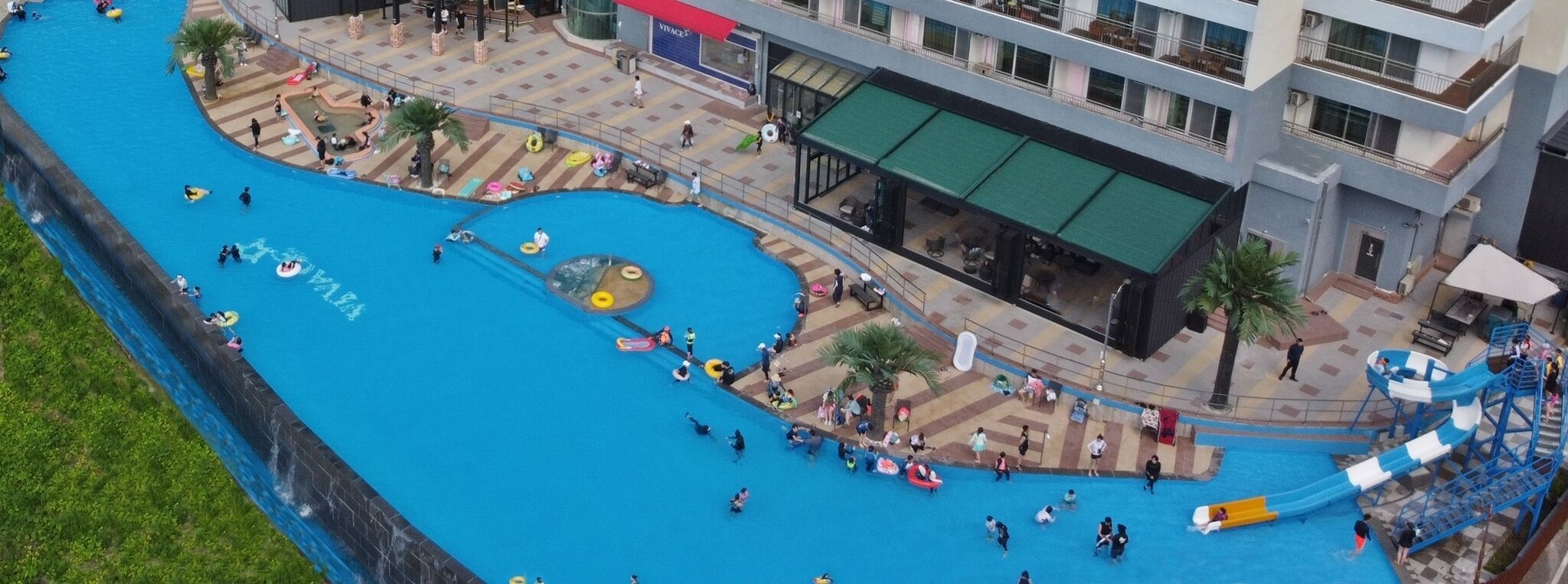 An aerial view of a public swimming area with multiple pools and slides, surrounded by buildings and outdoor seating areas.
