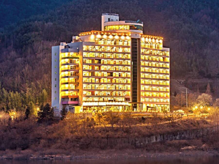 A multi-story hotel with a central atrium, surrounded by a body of water and forested hills.